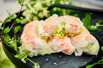 Colorful spring rolls filled with vegetables and shrimp are arranged on a dark, round tray. Lush green plants and delicate flowers surround the dish, creating a lively atmosphere.