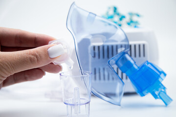 A close-up of a medicinal solution from an ampoule is dripped into an inhalation bowl against the background of a blue breathing mask and a device on a white background. The concept of treatment of