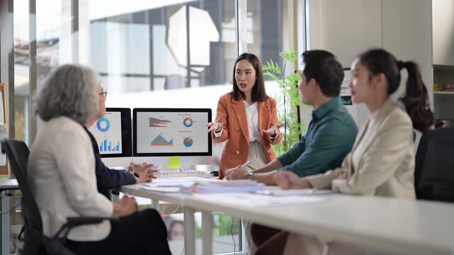 A group of people are sitting around a table with a woman pointing at a computer screen. The woman is wearing an orange jacket and is pointing at a graph on the screen