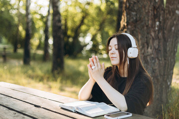 Young teen girl in headphones reading the Bible at a wooden table outdoors.