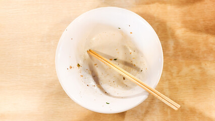 An empty white ceramic bowl with wooden chopsticks in it, displaying the remnants of a recently enjoyed meal of wonton noodles, signifies a satisfying culinary experience