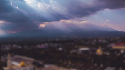 Dramatic sunlight streams through dark clouds over a mountain range and blurred cityscape below.