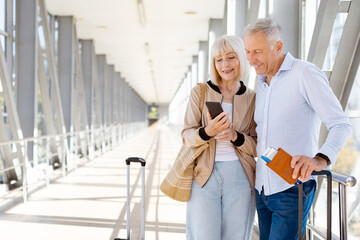 Senior couple using smartphone at airport while waiting with luggage and passports