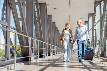 Happy senior couple walking at airport corridor with luggage, holding hands