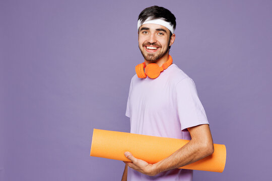 Young strong fitness trainer instructor sporty man sportsman wear headband t-shirt headphones train in home gym hold yoga mat carpet isolated on plain purple background. Workout sport fit abs concept.