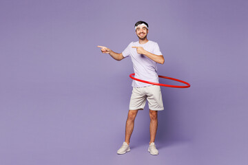 Full body young young strong fitness trainer instructor sporty man sportsman wear t-shirt train in home gym use hula hoop point aside isolated on plain purple background Workout sport fit abs concept