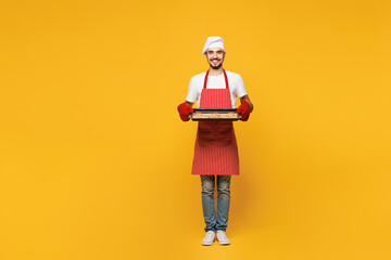 Full body young housewife housekeeper chef cook baker man wear red apron toque hat hold in hand tasty homemade chocolate cookies isolated on plain yellow orange background studio Cooking food concept