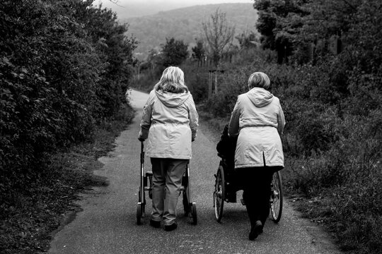 Elderly friends taking a walk in the park during cold weather with help of wheelchair and walker enjoying and having fun - Powered by Adobe