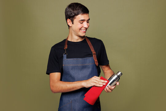 Young man barista bartender barman employee wear apron black t-shirt work in night club hold wiping shaker for preparing cocktails isolated on plain green background. Small business startup concept. - Powered by Adobe