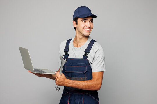 Young mechanic IT man wear overall hat uniform workwear clothes work in garage using laptop pc computer to diagnose car hold spanner isolated on plain grey background. Automotive repair job concept.