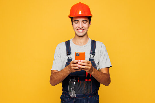 Young employee laborer handyman man he wear helmet overall hold use mobile cell phone isolated on plain yellow background. Instruments accessories for renovation apartment room. Repair home concept.