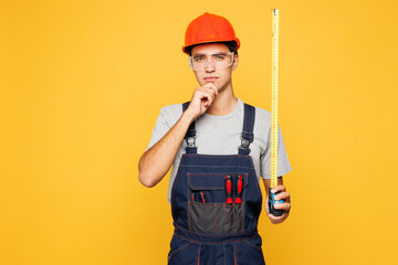 Young minded employee laborer handyman man wear helmet overall hold measuring tape think isolated...
