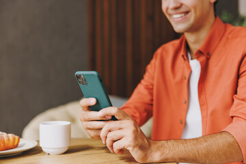 Cropped close up young man wear orange casual clothes use cell phone sitting alone at table in coffee shop cafe relax rest in restaurant in free time indoors. Freelance mobile office business concept.