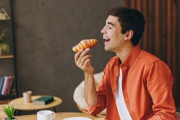 Side view young man wear orange casual clothes eat croissant bite sitting alone at table in coffee...