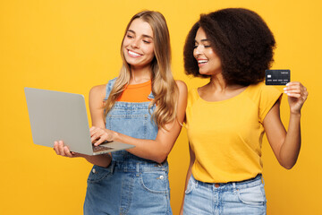 Two young friends buddies IT women wearing casual clothes together use laptop pc computer hold credit bank card shopping online order delivery booking tour isolated on plain yellow orange background