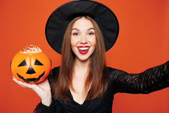 Fototapeta Close up young woman wears black hat dress witch costume doing selfie shot pov on mobile cell phone hold pumpkin isolated on plain red orange background. Celebration Halloween holiday party concept.