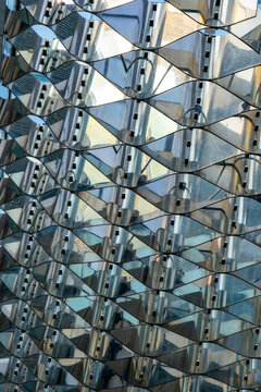 a modern architectural facade featuring a grid of irregular, diamond-shaped, reflective metallic panels. The panels create a dynamic, wave-like pattern, reflecting light and surrounding buildings
