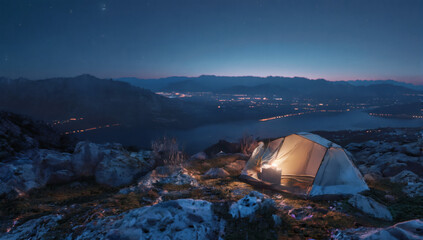 Glowing tent on a mountain overlooking city lights at blue hour. Atmospheric outdoor scene of a single illuminated camping tent on a grassy alpine slope above a valley of city lights at dusk. 