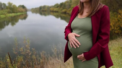 A pregnant woman in a green dress on the riverbank. A girl in a green dress. The expectant mother gently strokes her belly with her baby. Cloudy weather. Autumn park.