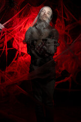Fototapeta premium Creepy man with scary Halloween zombie makeup trapped spider webs, trying to scare. Dead guy in cob web on red bloody background.