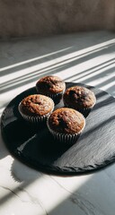 Four freshly baked muffins on a dark stone platter, illuminated by sunlight and shadow