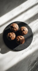 Four muffins arranged on a black slate, illuminated by striped sunlight over a marbled surface