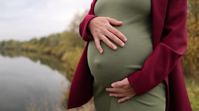 A pregnant woman in a green dress on the riverbank. A girl in a green dress. The expectant mother gently strokes her belly with her baby. Cloudy weather. Autumn park. - Powered by Adobe
