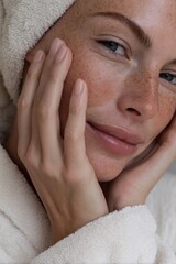 A close-up of a woman's face with visible freckles and soft fingers gently resting on her cheek while she wears a white towel robe and looks toward the camera