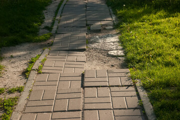 Broken tiled path leading into the distance with grass on both sides, parallel dirt trail on the left, deep shadows and a bright sunlit patch across the path.