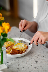 Woman cutting Croque Madame sandwich with knife and fork, served on white plate. Casual dining scene with yellow flowers, speckled countertop, and stylish black nail polish