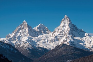 Triple snowy mountain summits under a clear blue sky; winter scenery featuring three peaks with snow and sunshine and forested base