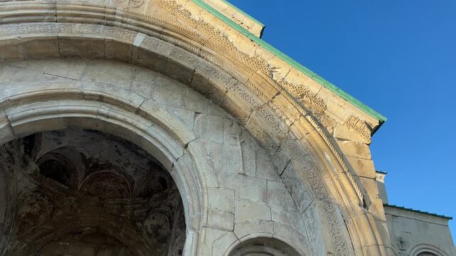 Close-up of ancient Georgian cathedral stone architecture: intricately carved arches and columns under a clear blue sky. Warm light accentuates its historical beauty and detailed craftsmanship.