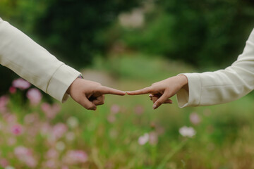 Two individuals reach out, their fingertips almost touching, against a soft focus natural backdrop filled with colorful flowers and lush greenery.