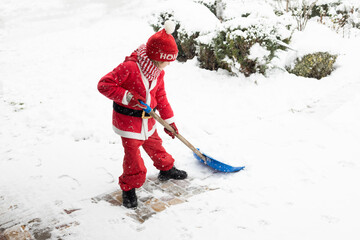 child in a red Santa suit with a large shovel clears snow in the yard after a snowstorm.