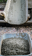A builder pours the remainder of wet concrete from a mixer truck into a wheelbarrow. Pouring cement mixture. Foundational work. Worker controls a mix flows through the gutter. Building industry.