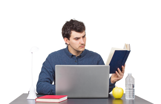 Focused young male student seated at the working desk in front of a laptop studying carefully from a book. Hard learning male looking for answers in a book, isolated portrait on transparent background