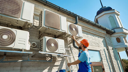 Technician inspecting outdoor air conditioning units under bright sunlight. Concept of engineering services and occupational safety for technical training, manuals, and professional publications.