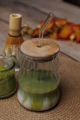 A glass of matcha tea latte with matcha powder in a glass jar and a matcha whisk on a braided mat.