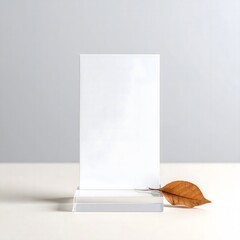 Minimalist blank white vertical card mockup on a transparent stand with a single dry autumn leaf, set against a soft neutral gray background