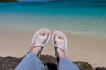 Feet with white sandals by emerald beach in Okinawa(沖縄の白砂ビーチと海を背景にした足元の写真）