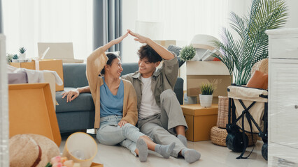 Fototapeta na wymiar Happy Asian couple sit on floor in front of sofa couch smile looking at camera making house sign in living room at new house. Husband and wife moving new house, Apartment Rent And Ownership Concept.