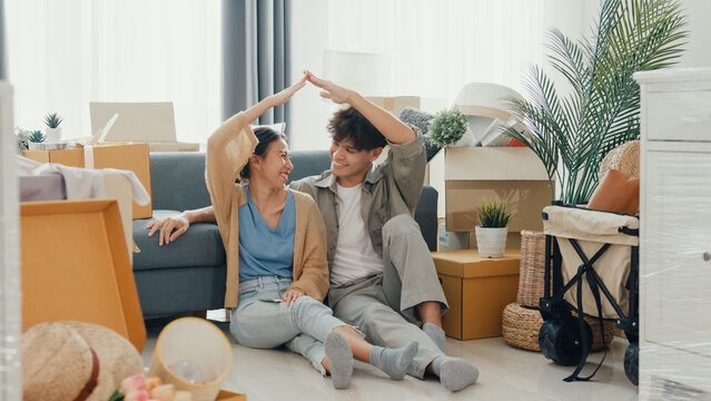 Happy Asian couple sit on floor in front of sofa couch smile looking at camera making house sign in living room at new house. Husband and wife moving new house, Apartment Rent And Ownership Concept. - Powered by Adobe
