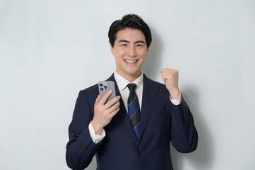 白背景でスマホを持ってガッツポーズをするスーツの日本人男性