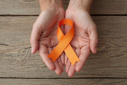 Close-up of hands clasping an orange awareness ribbon against a wooden surface, representing support for COPD, ADHD, leukemia, kidney and spinal cancers. Health and medical theme.