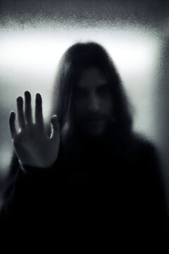 Mysterious figure with long hair reaching out through foggy glass in dimly lit setting during late evening