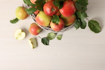 Fresh ripe apples with green leaves in wicker basket on white wooden table, top view. Space for text
