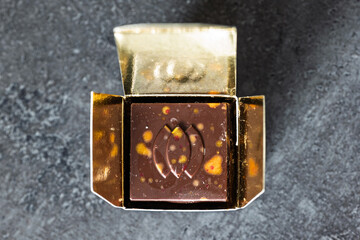 Single, premium, square chocolate praline in its open gold foil wrapper. Top down view, granite counter top background, close up, no people