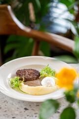 Gourmet dish with grilled meat patty, mashed potatoes, poached egg, brown gravy, and fresh greens on a white plate. Rustic table setting with flower and plants in background