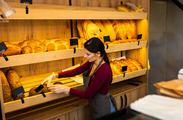 Smiling woman arranging freshly baked bread on shelves inside bakery.