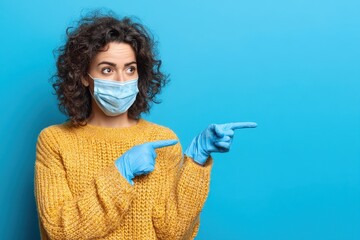 Mixed race young woman with brown hair wearing a mask and gloves gestures to the side while showcasing a product against a blue background